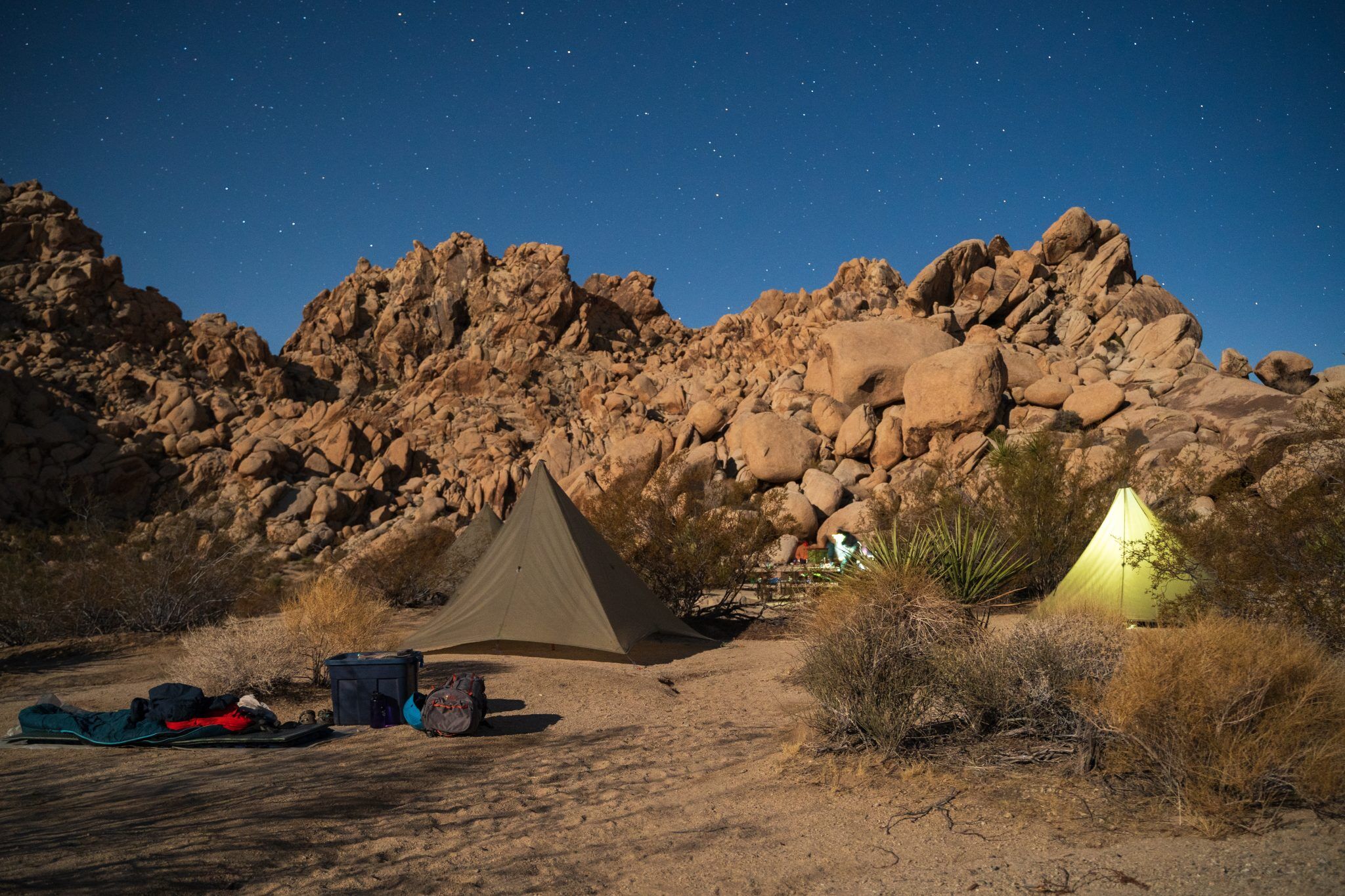 The image shows a campsite at night. There are two tents set up in a desert landscape with large rock formations in the background. Camping gear is scattered around the tents. The sky is dark and filled with stars, suggesting a clear night. The scene evokes a sense of tranquility and adventure.